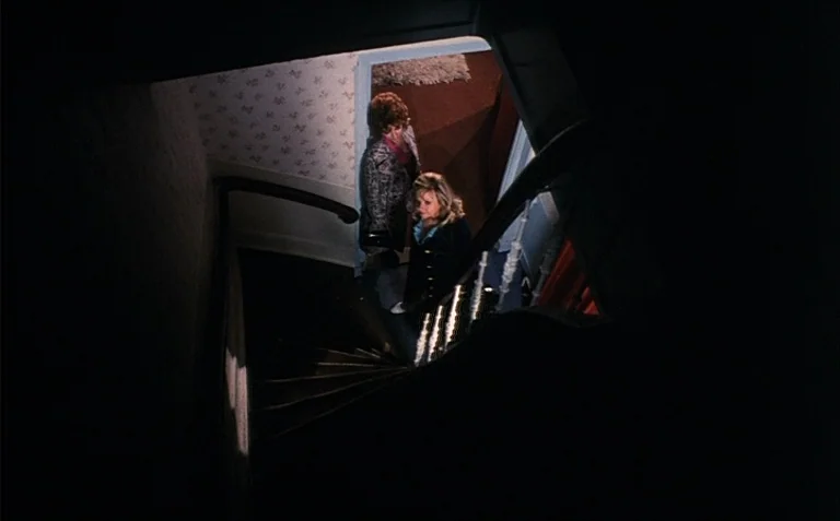 Business is Business (1971) movie still: wide — A dark stairwell featuring floral wallpaper and a wooden banister. Two women stand on the landing…