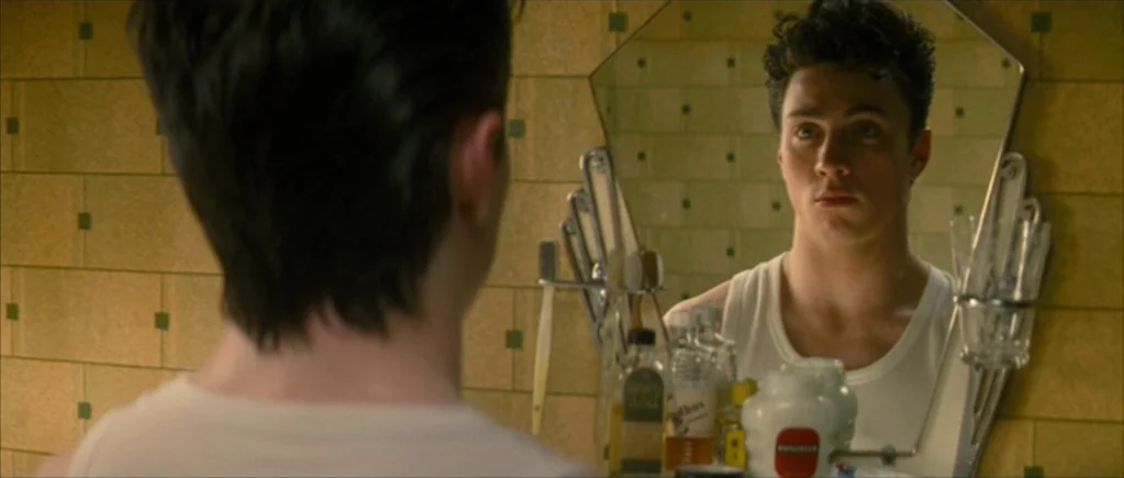 Nowhere Boy movie still: medium — A tiled bathroom features yellow wall tiles with small dark green inset squares. A young man stares intently at…