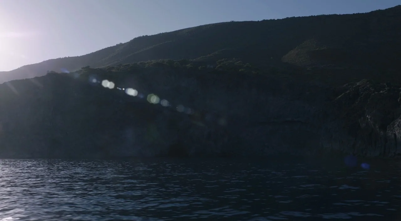 A Bigger Splash (2015) movie still: establishing — A dark rocky cliff rises sharply from the surface of a calm, deep sea. Sunlight flares across the…