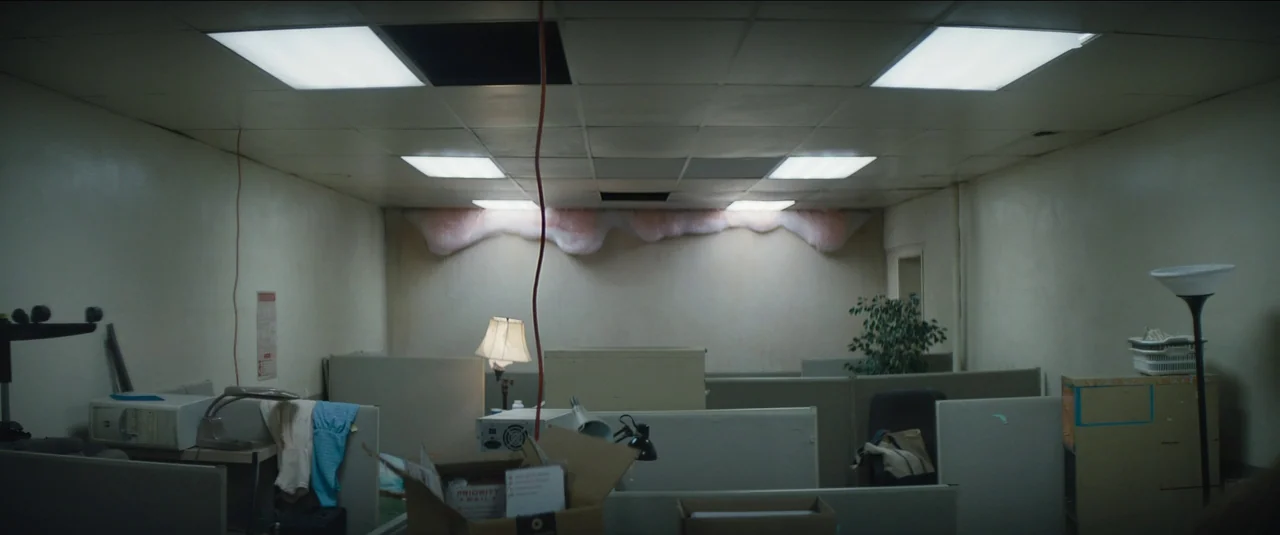 Kajillionaire (2020) movie still: establishing — A dimly lit, abandoned office space filled with low modular cubicle partitions. Pink insulation…