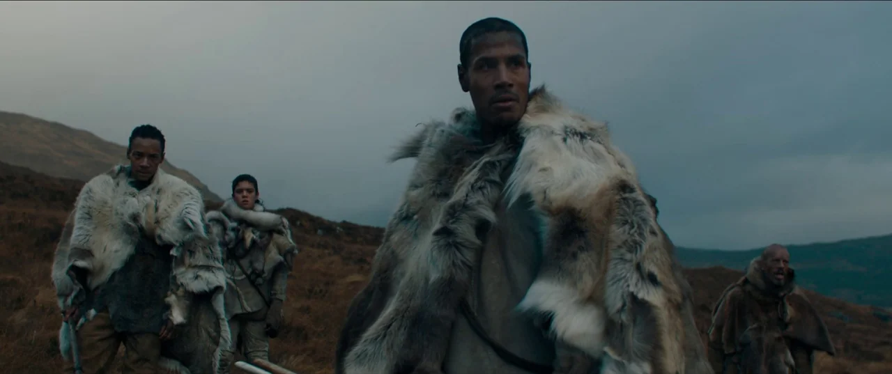 Out of Darkness (2022) movie still: medium — A group of four men walk across a barren, overcast moorland under a gray sky. The men wear thick furs…