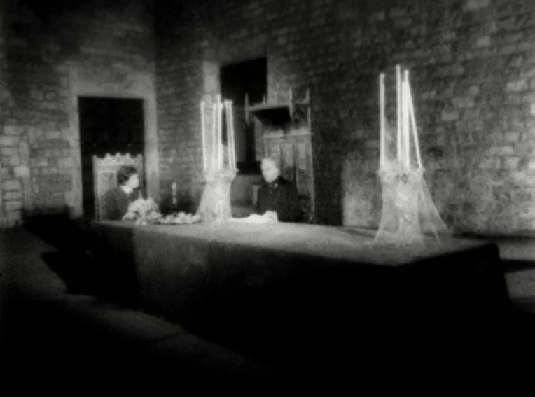 Cuadecuc, Vampir (1971) movie still: wide — A dark stone hall features a long table with two large candle holders. Two individuals are seated on…