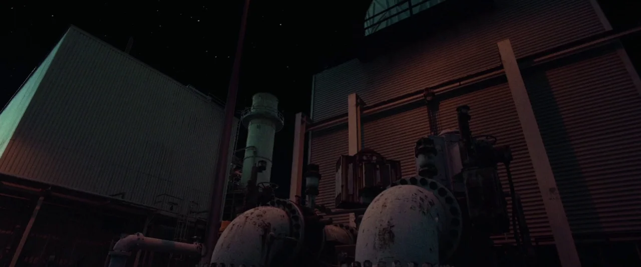 31 (2016) movie still: establishing — A dark, expansive industrial facility at night features large metal tanks and industrial pipes. The structure…