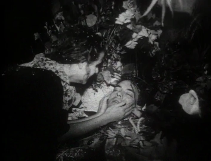 Archangel (1990) movie still: medium — A dimly lit interior space where a woman views a man in an open casket. She holds the face of the deceased…