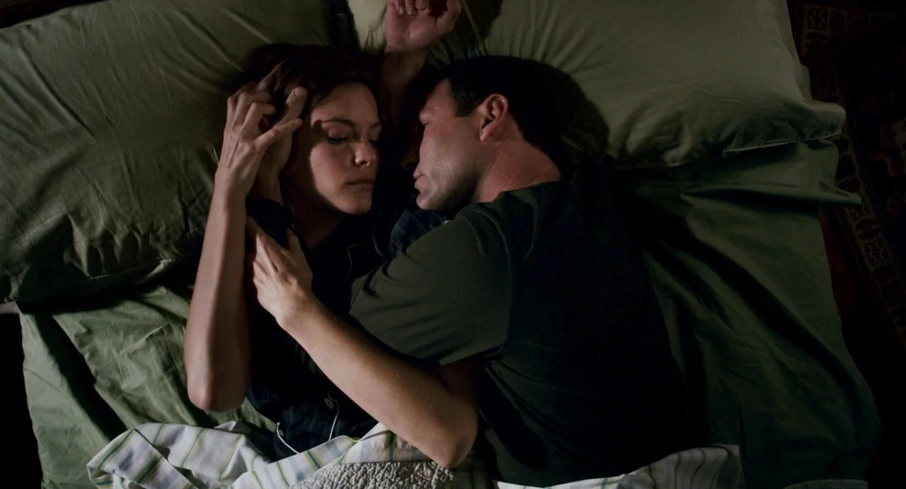 Super (2010) movie still: medium — A dark, olive-toned bed features two people resting with their eyes closed. The man holds the woman close while…