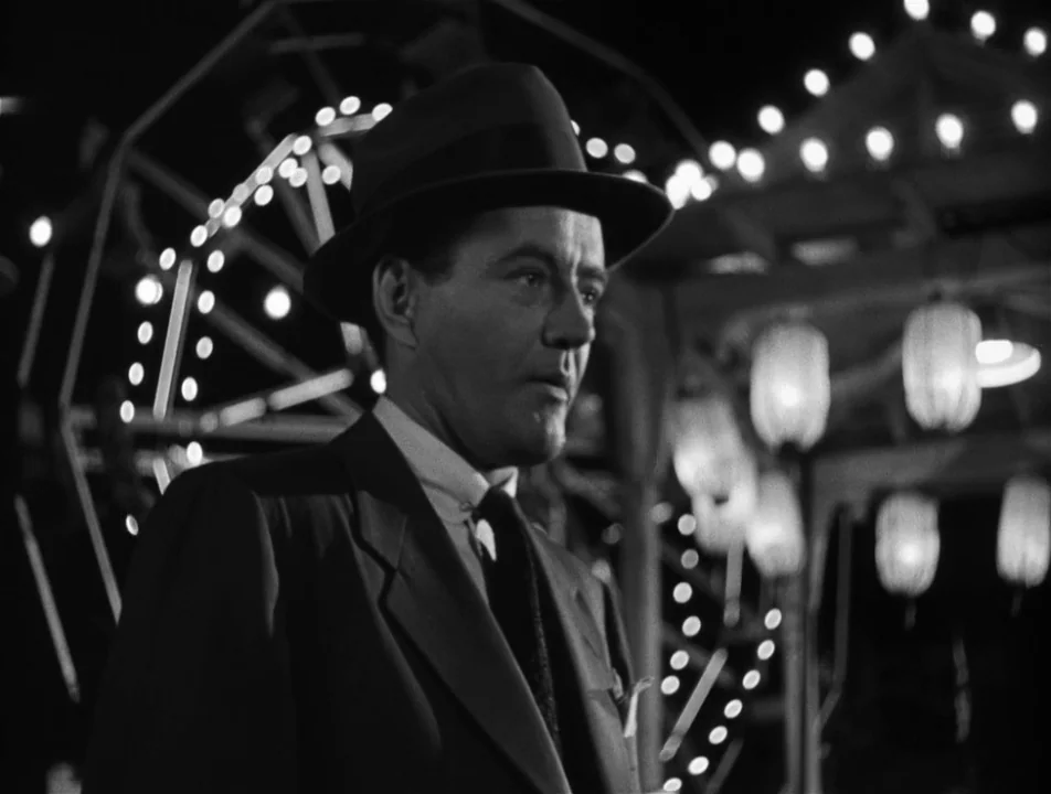 Strangers on a Train (1951) movie still: medium — A night scene at a fairground featuring blurred rides and glowing decorative light bulbs. A man in…