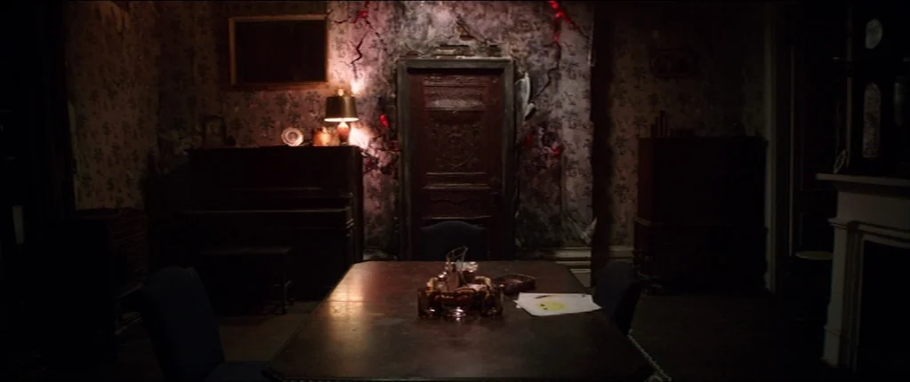 Abattoir (2016) movie still: wide — A dark, dilapidated dining room features crumbling wallpaper and a heavy, ornate wooden door. A set dining table…
