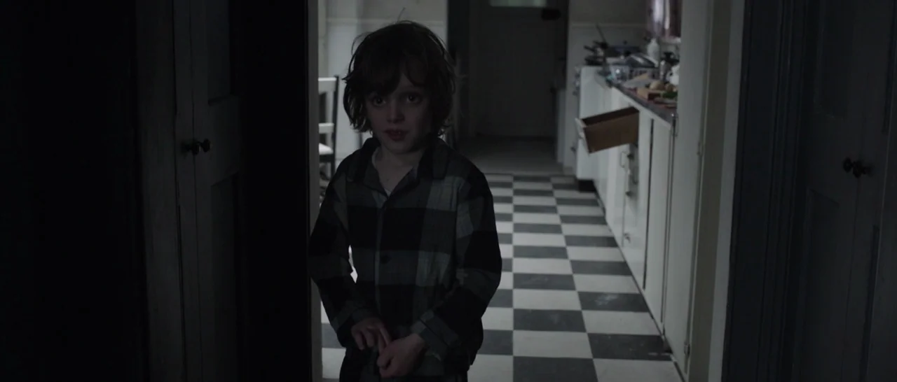 The Babadook (2014) movie still: medium — A dimly lit doorway opens into a room with a black and white checkered floor. A young boy in plaid pajamas…