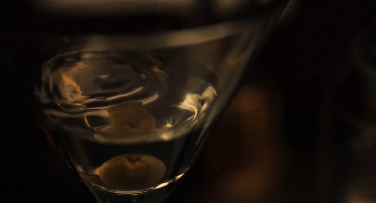 On The Rocks (2020) movie still: closeup — A close-up view of a glass containing a submerged olive in a dark environment. The liquid surface…