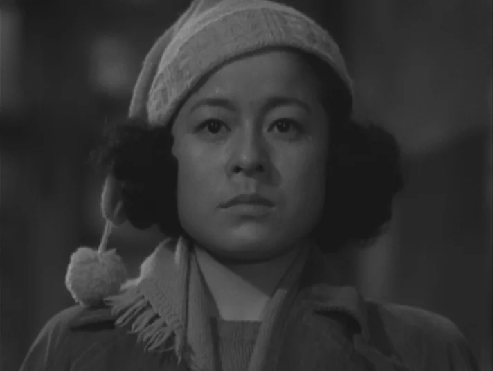 Scandal (1950) movie still: closeup — A woman stands indoors against an indistinct, dimly lit background. She looks directly toward the camera…