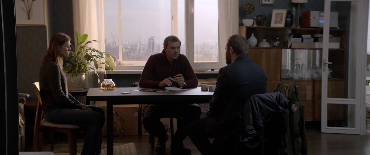Loveless (2017) movie still: wide — An apartment interior features a dining table placed before a large window with city views. Three people sit…