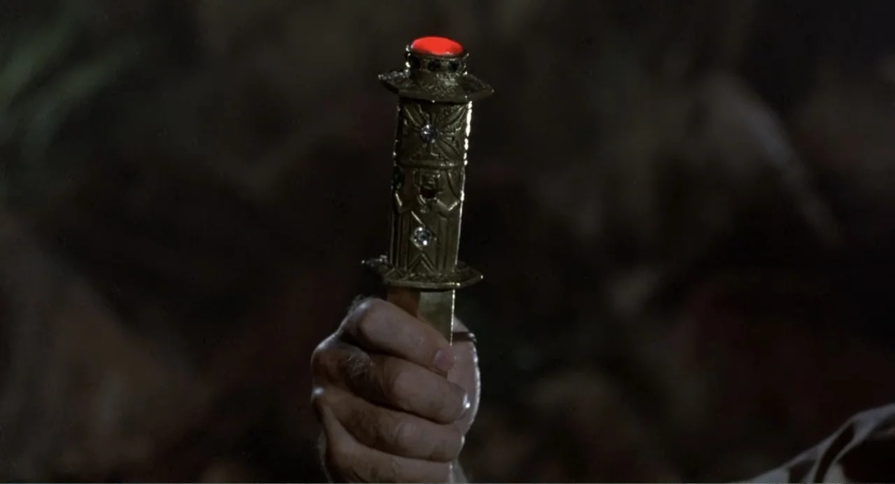 Firewalker (1986) movie still: closeup — A dark, rocky environment serves as the background for a held ceremonial blade. A hand firmly grips the…