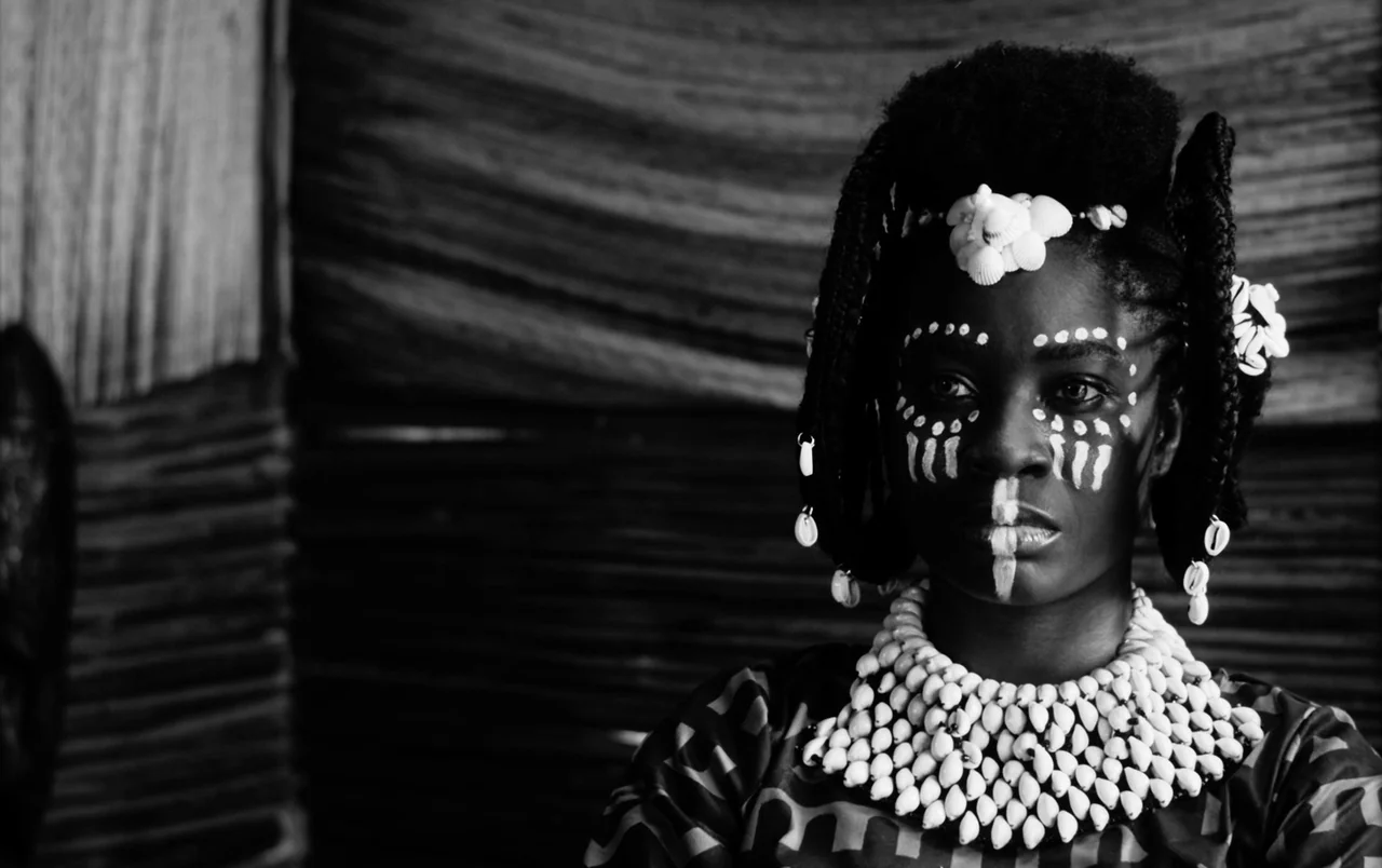 Mami Wata (2023) movie still: closeup — A woman stands in a dimly lit space with a textured woven wall behind her. She wears traditional shell…
