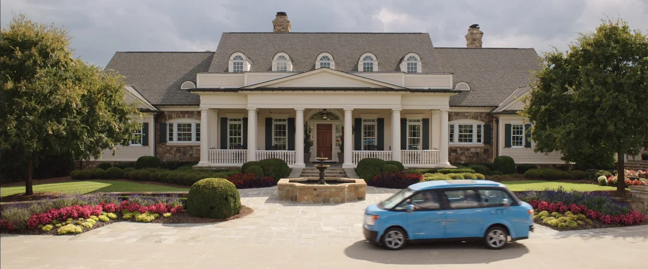 Vacation (2015) movie still: wide — A large multi-story suburban mansion features white columns and stone masonry accents. A light blue minivan…