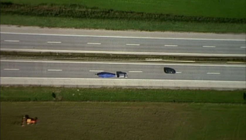 Funny Games (1997) movie still: establishing — An overhead view of a two-lane highway cutting through wide green agricultural fields. A truck with a…