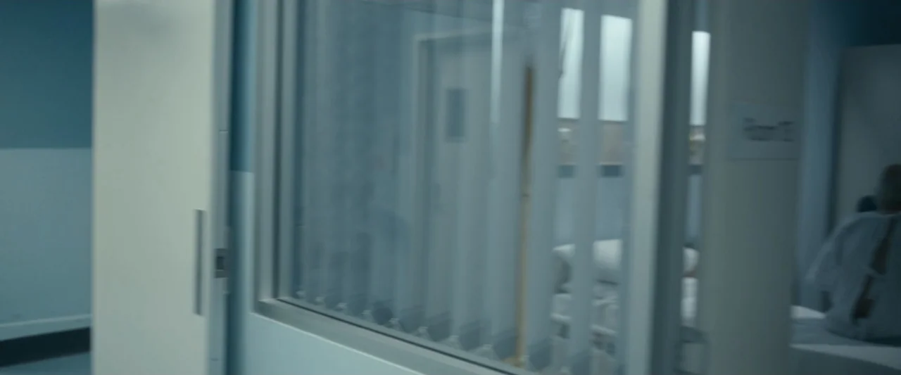 Talk to Me (2023) movie still: medium — A sterile hospital room features blue-painted walls and a glass panel partition with vertical blinds. A…