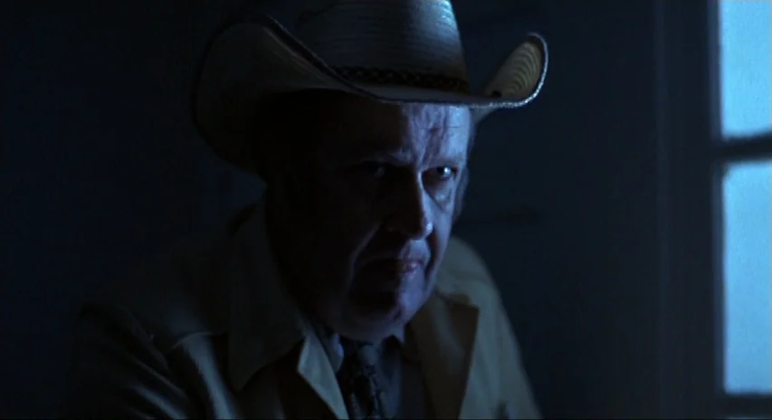 Blood Simple (1984) movie still: closeup — A dimly lit room with a man positioned near a window at night. The man wearing a cowboy hat stares…
