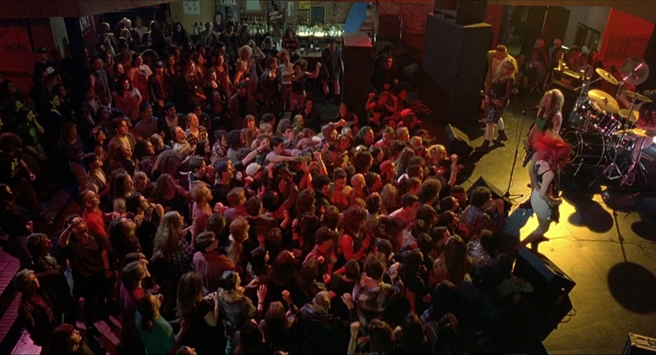 Serial Mom (1994) movie still: wide — A dense crowd of people occupies a dark room facing a small stage area. Musicians perform with guitars and…