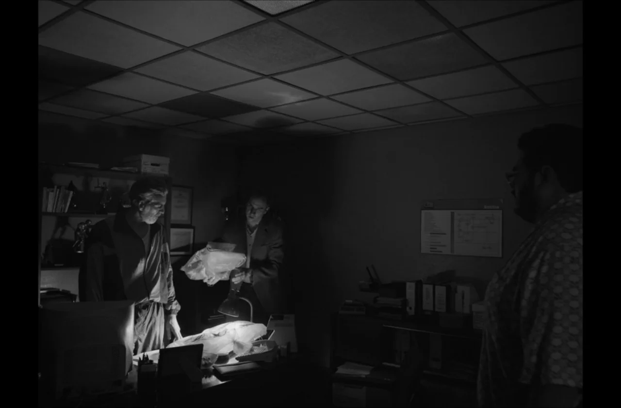 The Kitchen (2012) movie still: medium — A dim office space contains a desk occupied by three men under artificial light. Two men inspect a clear…