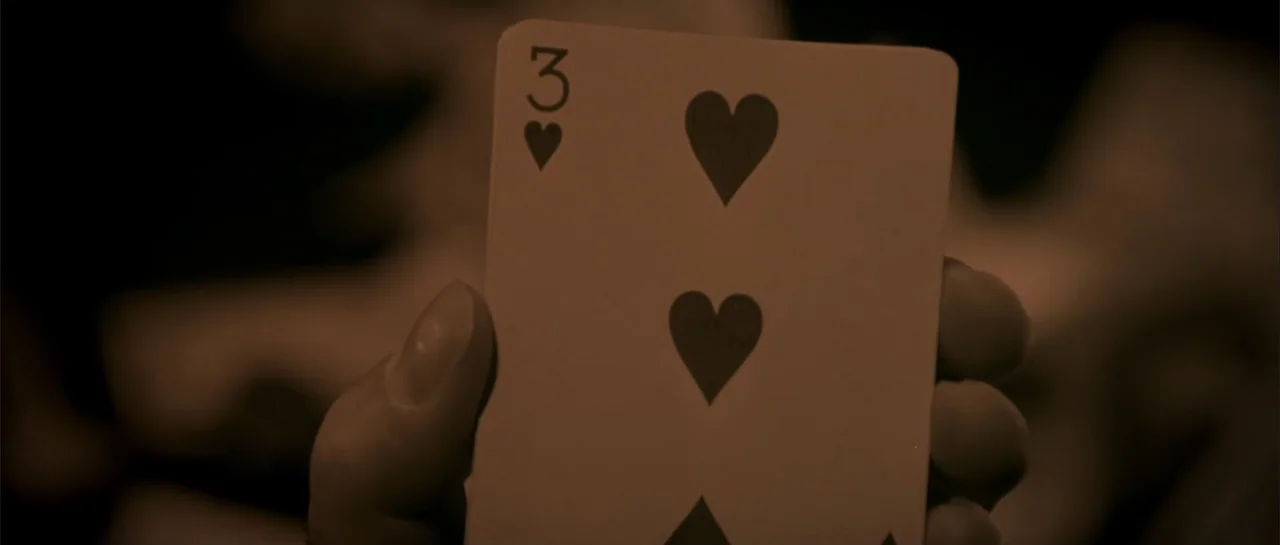 Shade (2003) movie still: closeup — A dimly lit interior space features an out-of-focus background with deep shadows. A human hand holds up a single…
