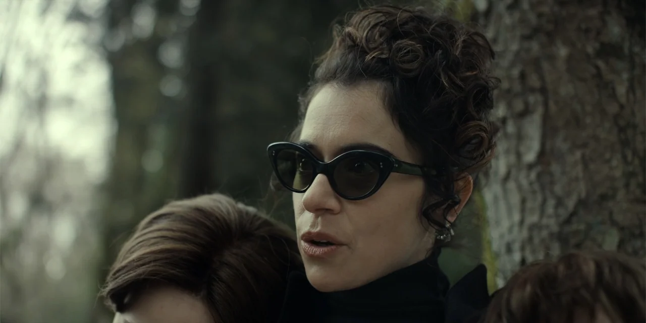 The Monkey (2025) movie still: closeup — A woman stands in a dense woodland setting with textured bark visible behind her. She wears black cat-eye…