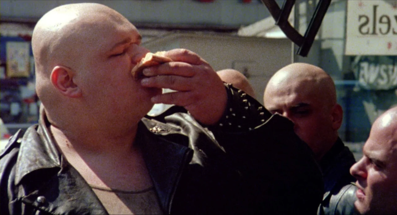 The Wanderers (1979) movie still: medium — An outdoor urban environment featuring a group of men wearing black leather jackets. One bald man stands…