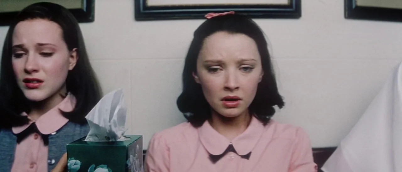 Pretty Persuasion (2005) movie still: medium — Two women wearing identical pink collared shirts sit together in a light-walled room. A box of…