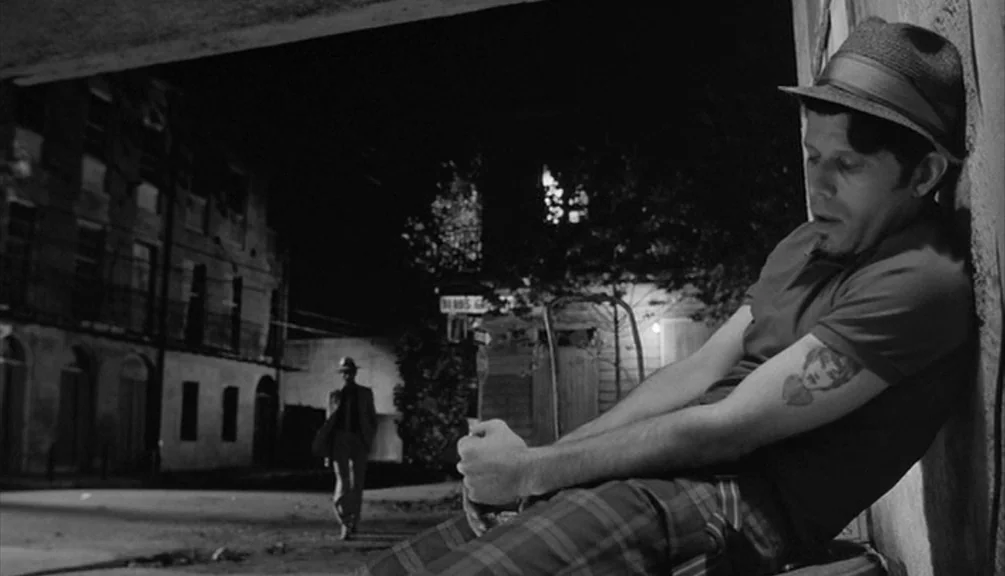 Down By Law (1986) movie still: medium — A night scene shows a dimly lit urban alleyway with dark, textured architecture. One man sits hunched…
