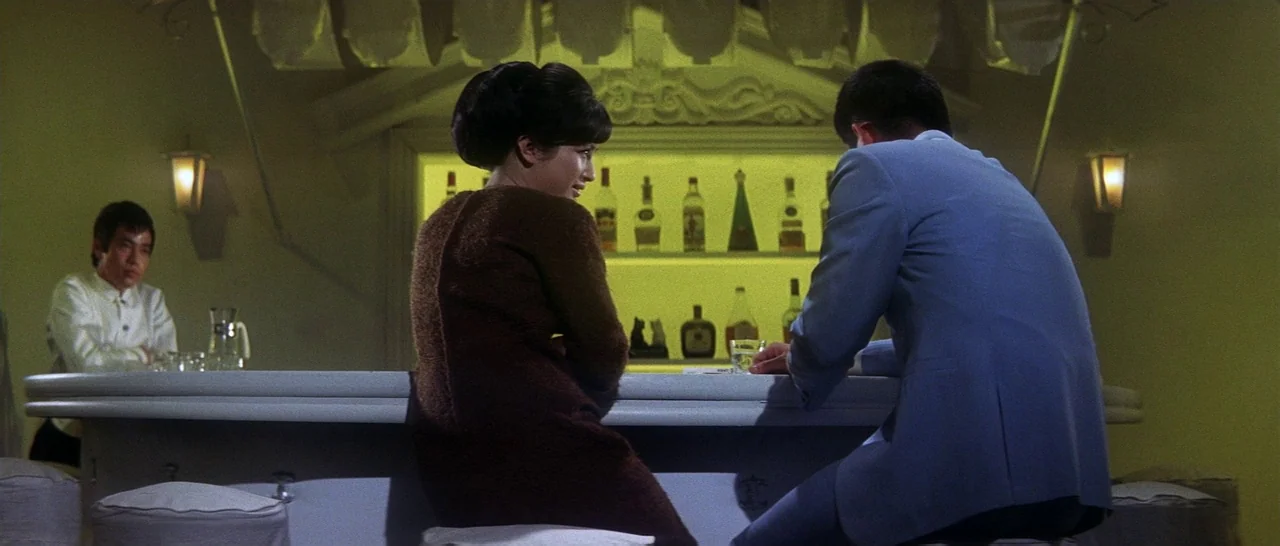 Tokyo Drifter (1966) movie still: medium — A dimly lit mid-century bar features rows of liquor bottles on shelves. A woman and man sit at the…