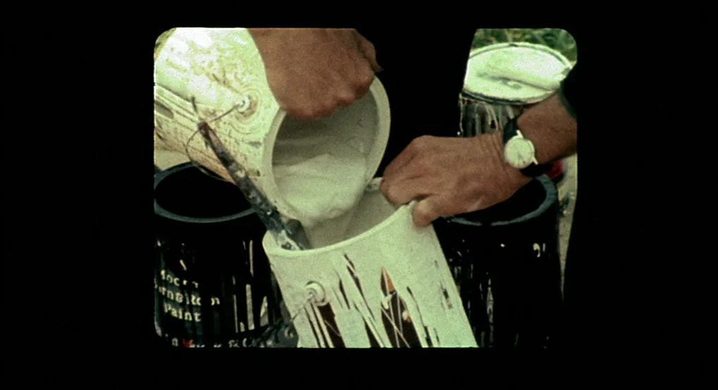 Pollock (2000) movie still: closeup — Two hands pour thick white paint between old, dented metal cans. The containers are stained with remnants of…