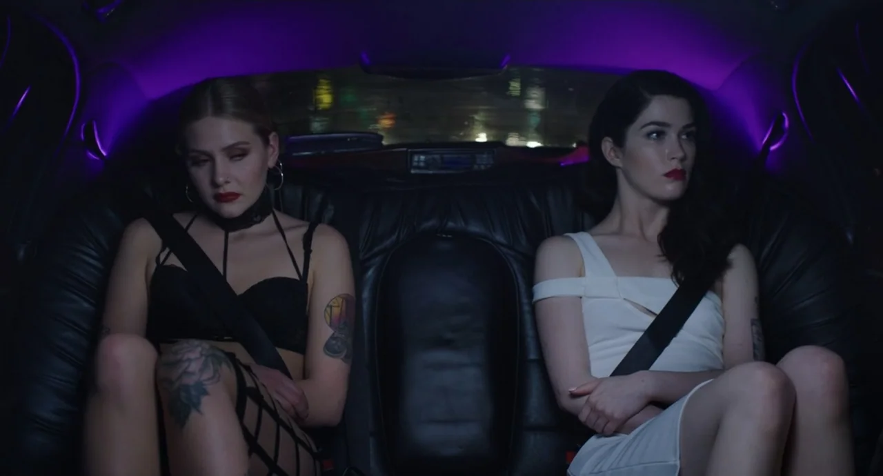 Pleasure (2021) movie still: medium — Two women sit in the leather backseat of a dark vehicle at night. The interior is bathed in purple accent…