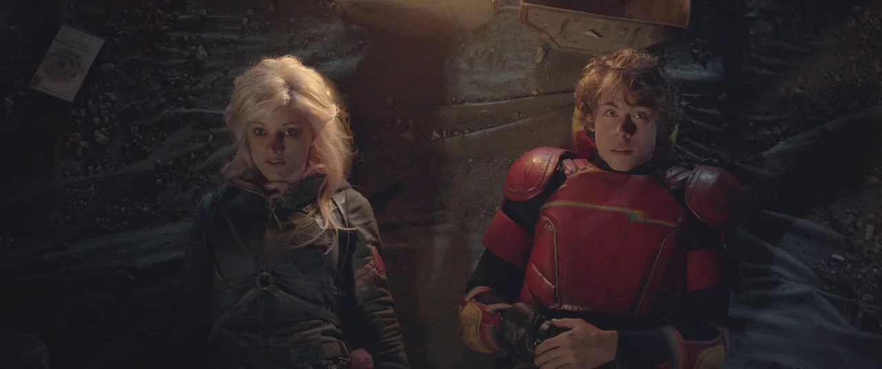 Turbo Kid (2015) movie still: medium — A dark, uneven surface covered in rocky debris and scattered industrial waste components. Two individuals in…