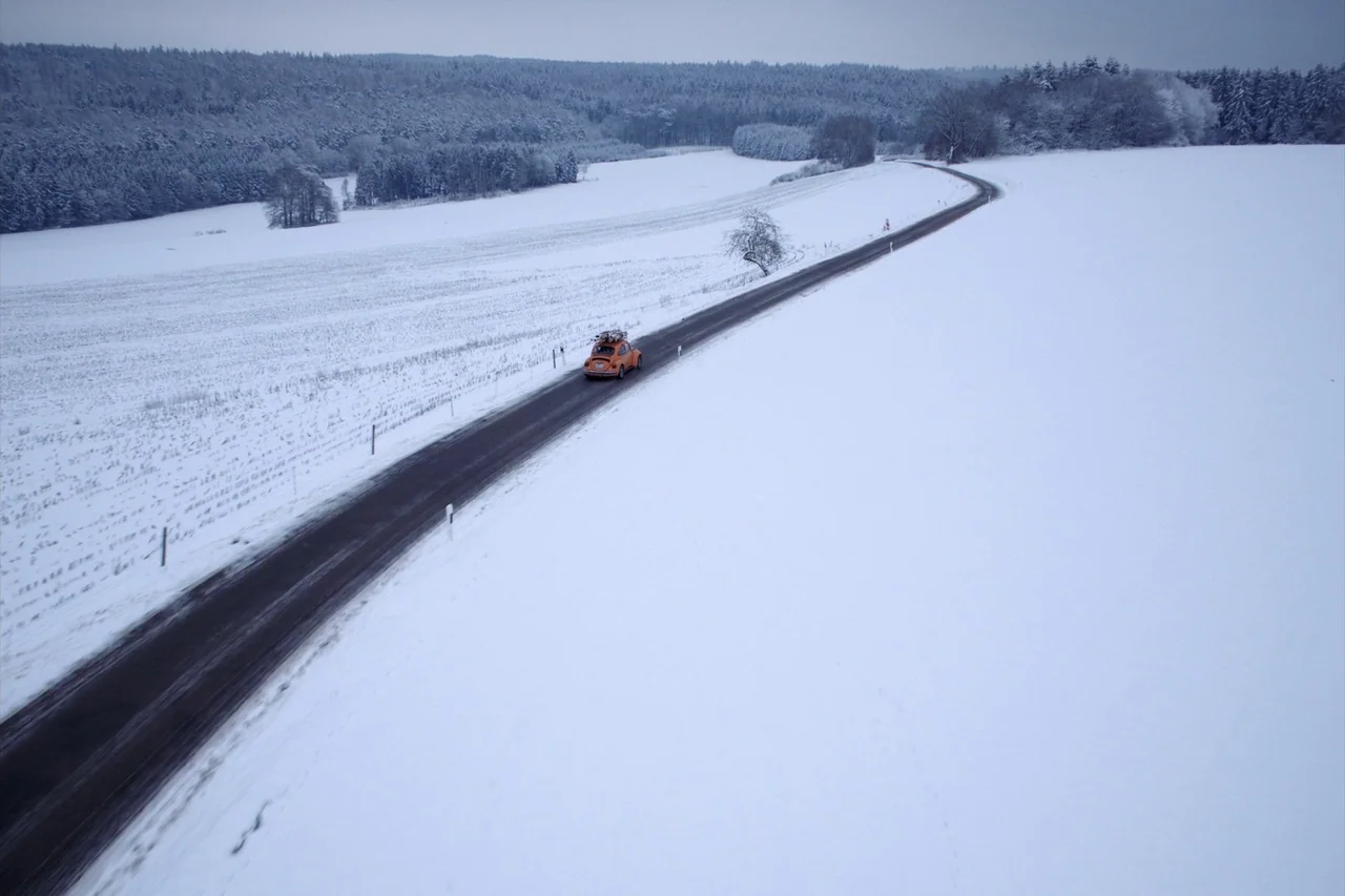 Anselm (2023) movie still: wide — A snow-covered rural landscape features a single asphalt road cutting through fields and trees. An orange vintage…