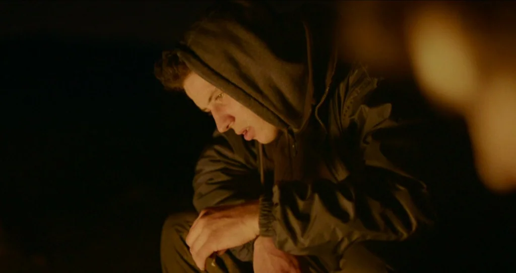 God’s Own Country (2017) movie still: medium — An outdoor setting at night illuminated by a nearby fire source. A young man wearing a hoodie sits…