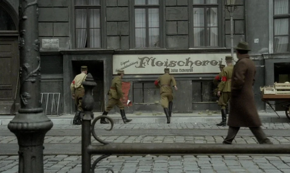 Despair (1978) movie still: wide — A cobblestone street in front of a German butcher shop with stone architecture. Several soldiers in uniform…