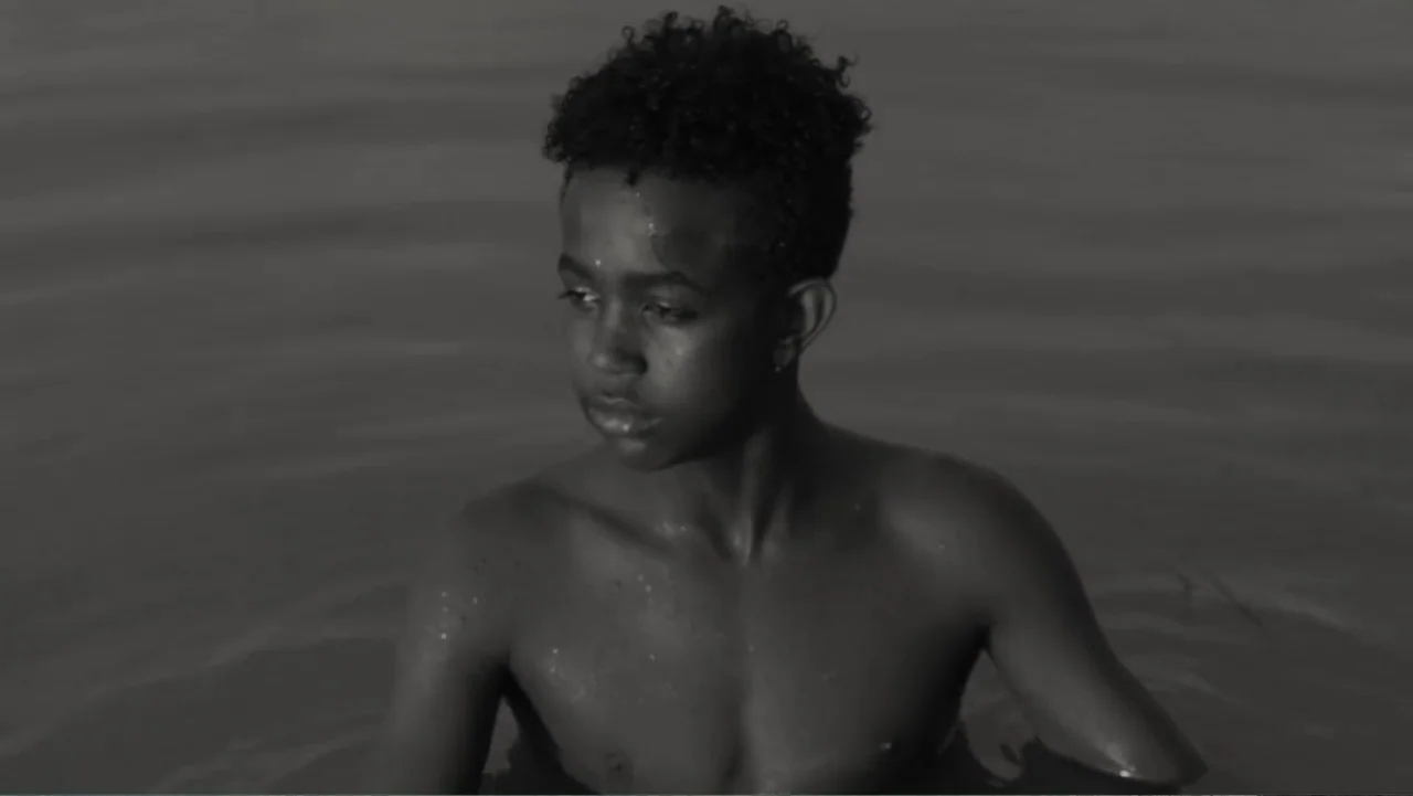 Faya Dayi (2021) movie still: medium — A shirtless adolescent boy stands neck-deep in calm, dark water. The boy is looking off-camera to his left…