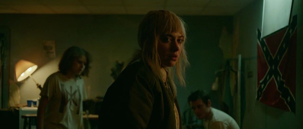 Green Room (2015) movie still: medium — A dark, dimly lit room contains three people with a Confederate flag mounted on the wall. A blonde woman…