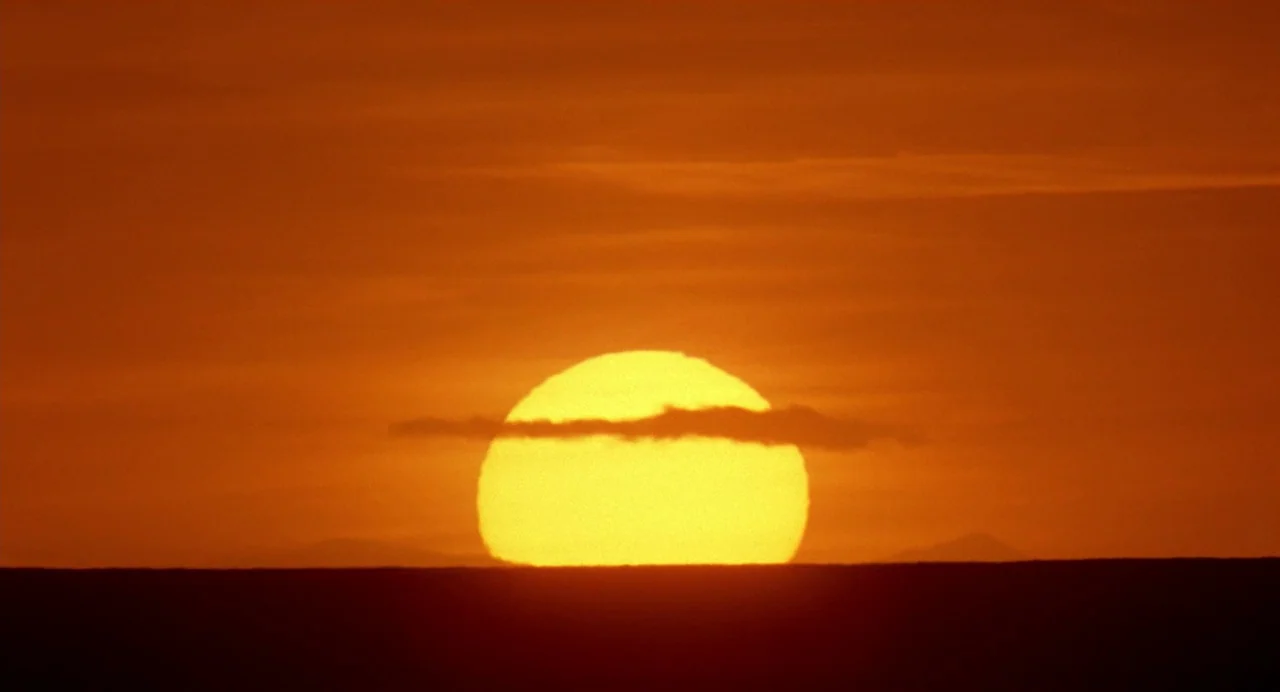 The Reflecting Skin (1990) movie still: establishing — The sun descends towards the flat line of the horizon during sunset. A thin layer of clouds…