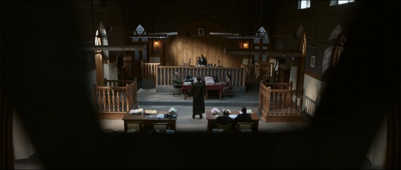 Garudan (2024) movie still: wide — A quiet courtroom features wooden paneling and a raised judge bench with numerous law books. A single lawyer…