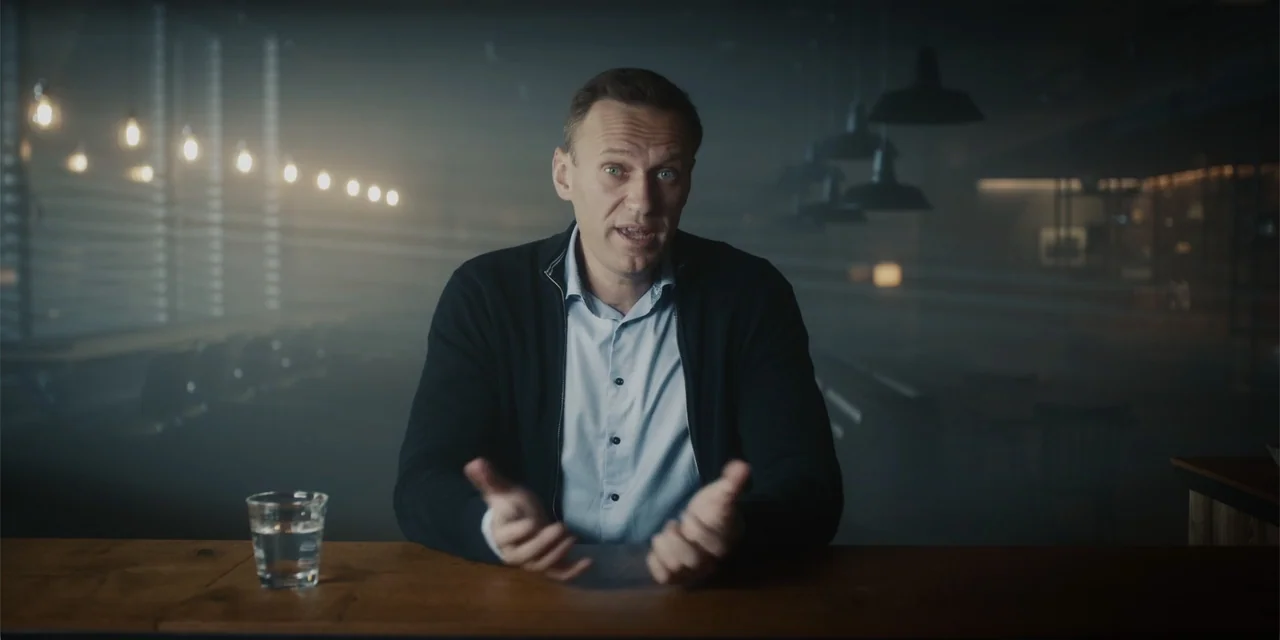 Navalny (2022) movie still: medium — A dimly lit room with a wooden bar counter and scattered industrial light fixtures. A middle-aged man sits…