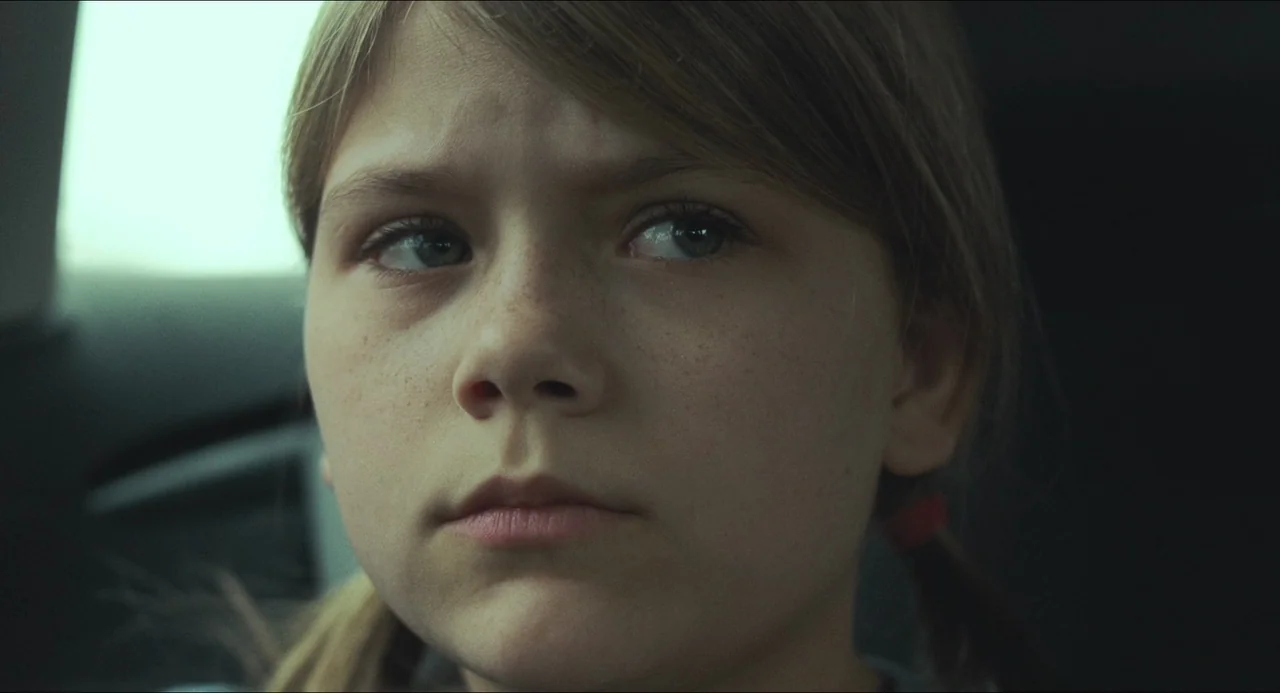 The Lodge (2019) movie still: closeup — The interior of a car during the day with light filtering through the window. A young girl sits in the…