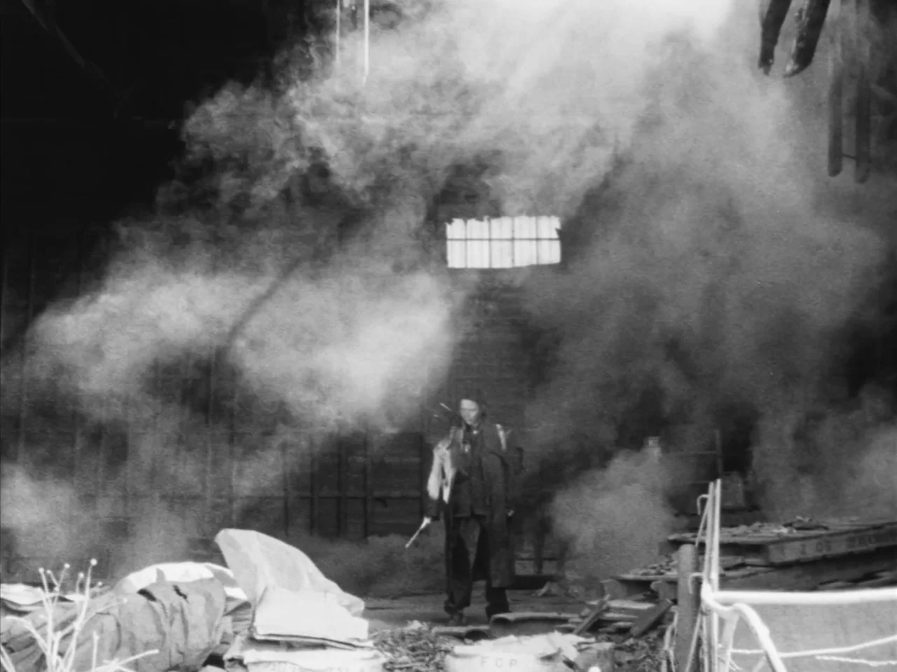 Tetsuo: The Iron Man (1989) movie still: wide — A dark, dilapidated industrial space is filled with dense swirling smoke and debris. A single figure…