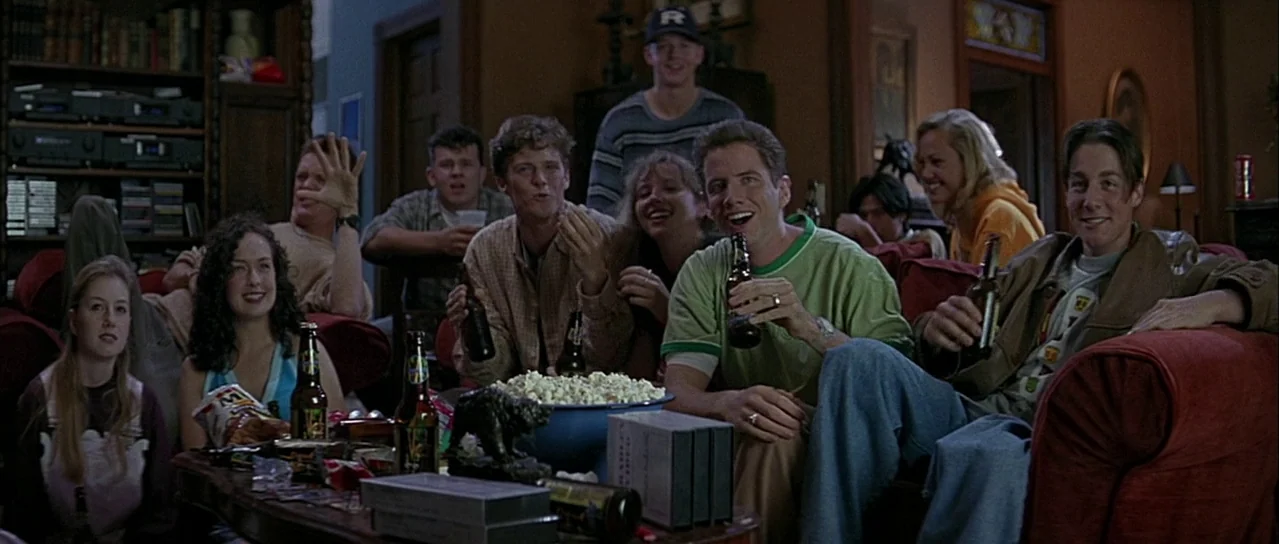 Scream (1996) movie still: wide — A group of young adults congregates on red sofas in a 1990s domestic space. The group watches television while…