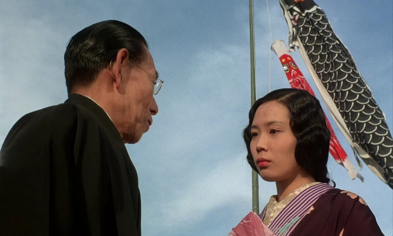 In The Realm of the Senses (1976) movie still: medium — An outdoor space features a flagpole with a decorative carp streamer against a sky. A man in…