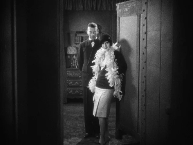 Underworld (1927) movie still: fullbody — A 1920s interior space features a dark wood doorway with a heavy metal panel. A woman wearing a feathered…