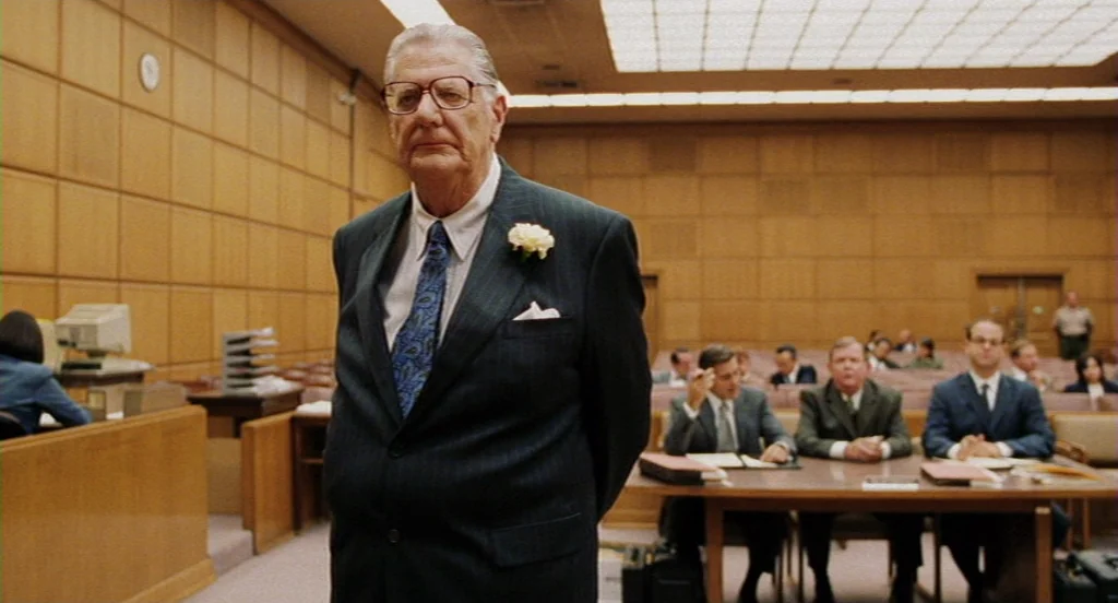 Intolerable Cruelty (2003) movie still: medium — A wood-paneled room features light-colored walls and rows of seated men in suits. An elderly man in…