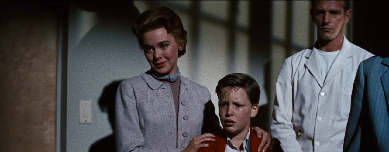 Bigger Than Life (1956) movie still: medium — A dimly lit indoor space features a wall with visible window-pane shadow patterns. A woman…
