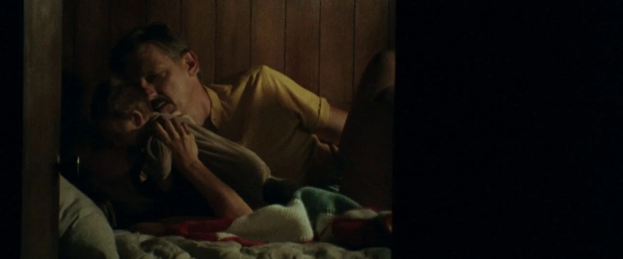 Adoration (2019) movie still: medium — A dark bedroom features wood-paneled walls and a bed with a colorful knitted blanket. An adult man lies down…
