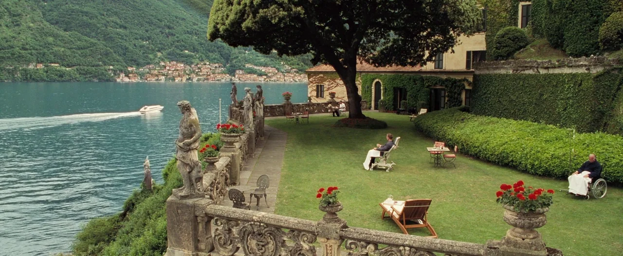 Casino Royale (2006) movie still: wide — A stone-walled garden terrace overlooks a large lake surrounded by forested mountains. Two men sit on…
