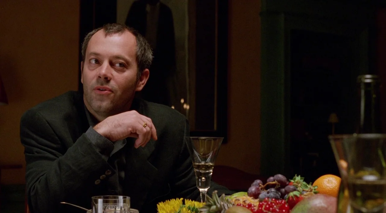 Shallow Grave (1994) movie still: medium — A dimly lit room features a table set with bowls of fresh fruit and wine glasses. A middle-aged man in a…